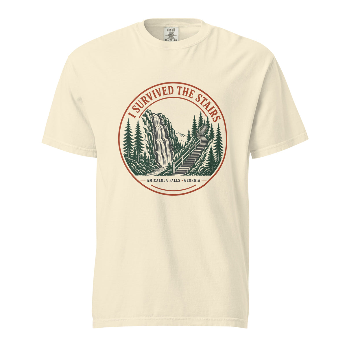 Ivory Comfort Colors tee, flat front view, with a round red and green 'I Survived the Stairs' badge showing Amicalola Falls and the trail staircase through pine trees